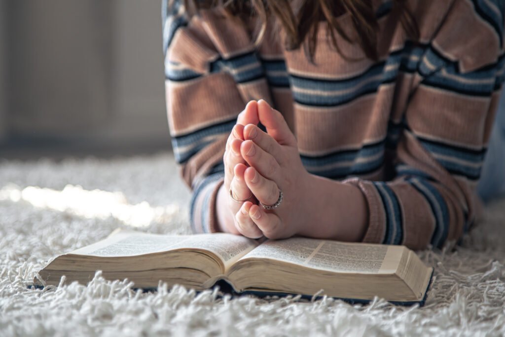 A close-up of a young woman with a striped sweater, lying on the floor, holding her hands together in prayer over an open Bible.