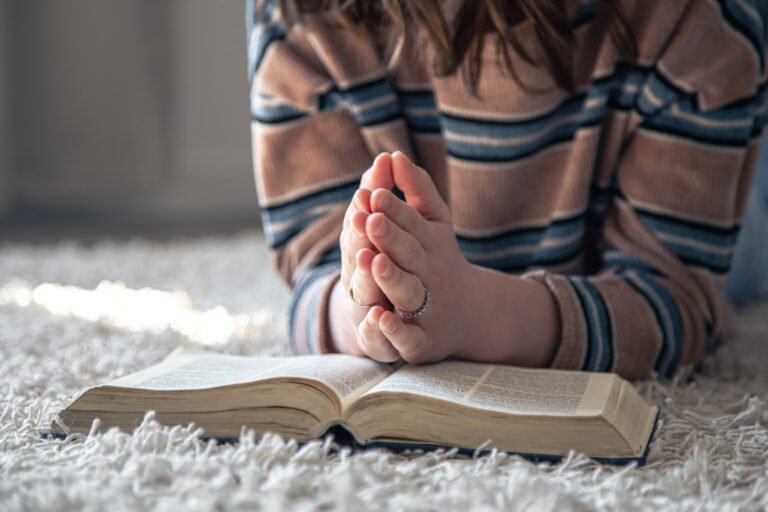 A close-up of a young woman with a striped sweater, lying on the floor, holding her hands together in prayer over an open Bible.