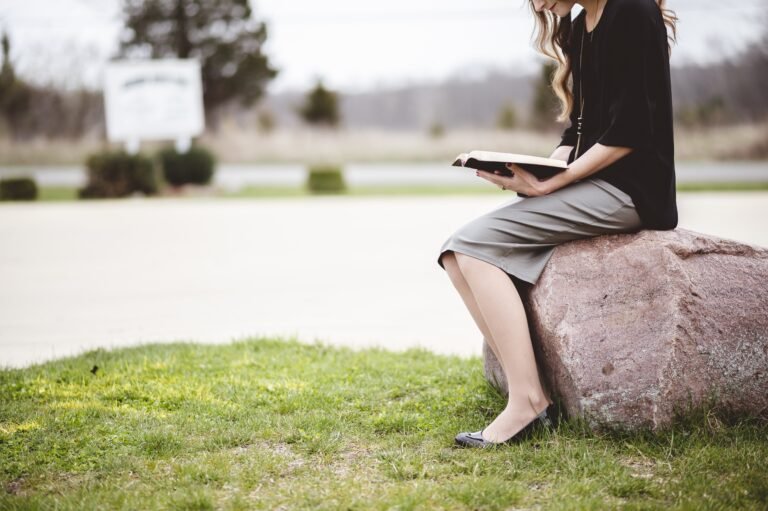 A woman sitting on a large rock, reading a book, wearing a black top and grey skirt.